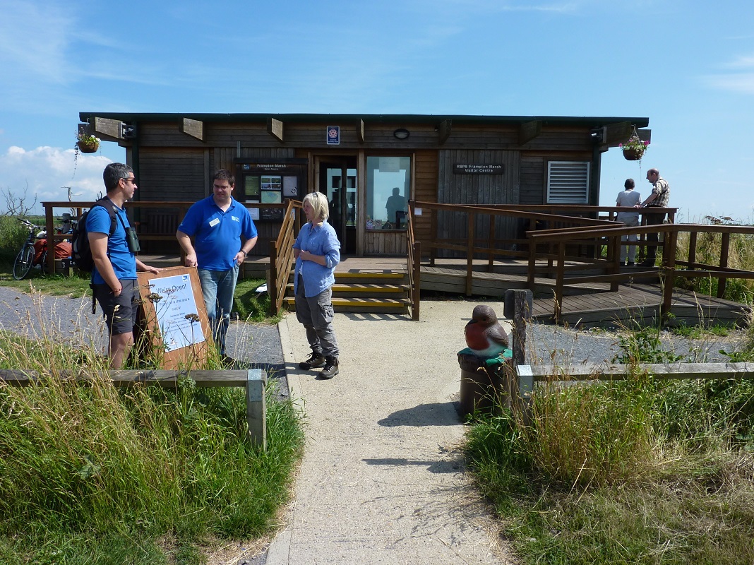 A colour photo of Frampton Marsh Visitor Centre. A male and female member of staff are talking to a member of the public. Behind them is the wooden visitor centre which is surrounded by wild land. Two people are stood beside the building looking out across the marsh.