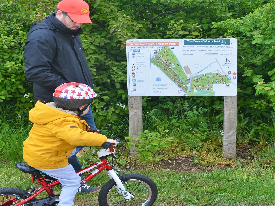 A man and his child, riding a bicycle, at a map board. The map depicts the area of Sir Joseph Banks country park