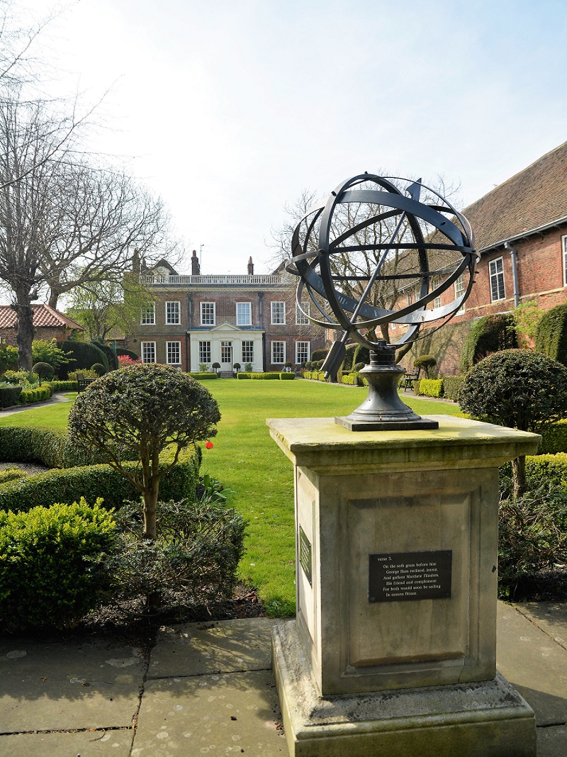 A photograph Fydell House garden. In the foreground is a large Armillary Sphere on a plinth. The walled garden consists of a large lawn surrounded by a path and low sculpted hedges. In the background is the rear of Fydell House.