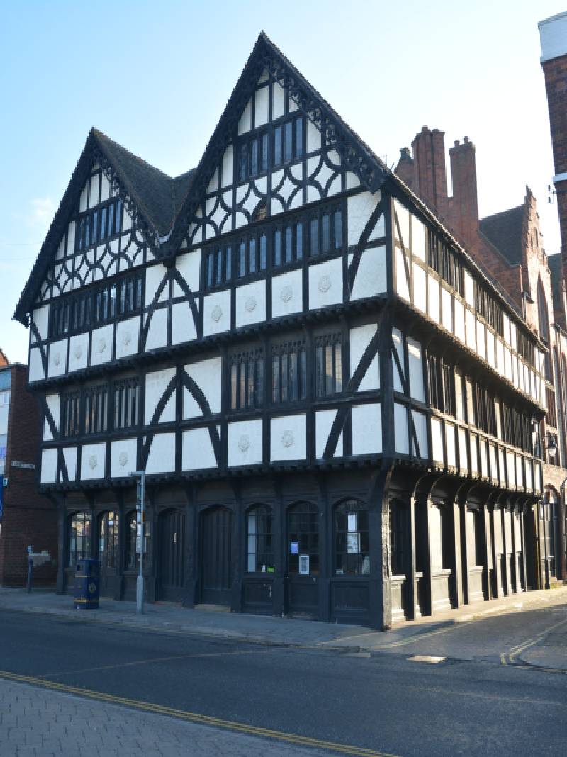 A coloured of Shodfriars Hall in the Year 2018 showing the original features. The wooden frame remains, as well as the windows and roof decorations. The wooden panels at the base of the building as well as the frame are stained black, and the daub between the wooden frames is white.