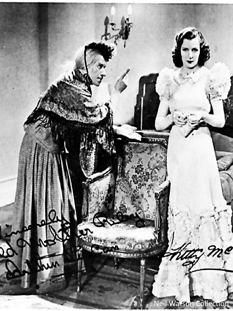 A black and white photo of the production of Mother Riley. A male actor is dressed as a washerwoman, pointing at a female actress, his wife, playing the young daughter. The actress wears a floor length dressed with a frilled hem and large frilled sleeves. The male actor is wearing a long skirt and jacket, as well as shawl, wig and cap. The photograph is signed by both actors with a message which is illegible.